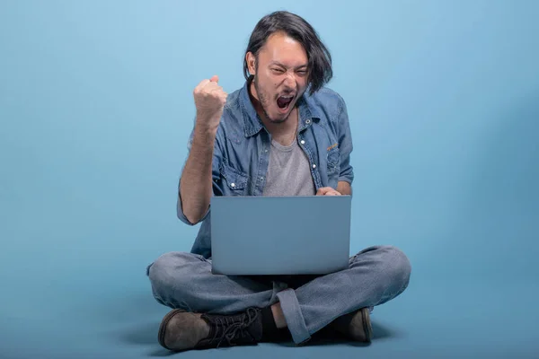 Young bearded man sit down on floor using computer in blue background. Excited Asian young hipster using laptop, full body shot. Young generation hipster working concept.