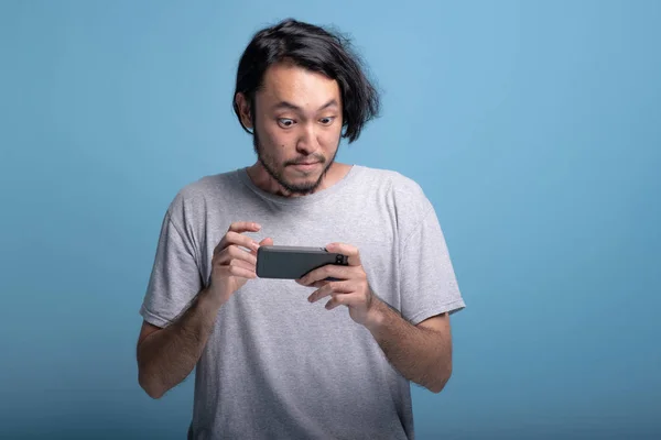 Excited young bearded man playing mobile games in blue background. Extremely surprise Asian young hipster playing mobile game, half body shot. Young generation hipster relaxing concept.