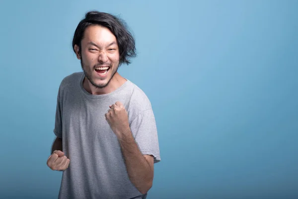Young bearded man winning pose in blue background. Happy Asian young hipster closed fist winner pose, half body shot. Young generation hipster concept.