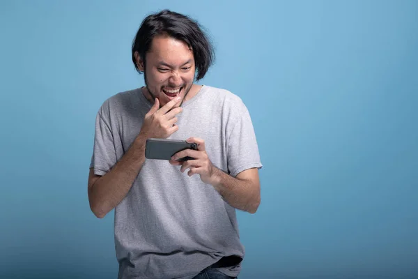 Excited young bearded man playing mobile games in blue background. Extremely excited Asian young hipster playing mobile game, half body shot. Young generation hipster relaxing concept.