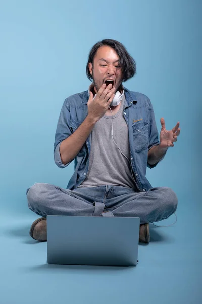 Young bearded man sit down on floor using computer in blue background. Shocked Asian young hipster using laptop, full body shot. Young generation hipster working concept.