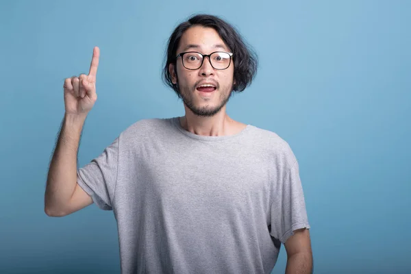 Young bearded man pointing finger in blue background. Excited Asian young hipster pointing finger upwards, half body shot. Young generation hipster idea concept.