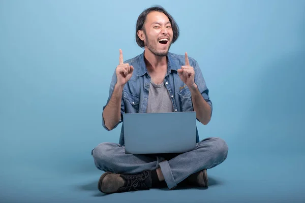 Young bearded man sit down on floor using computer in blue background. Very excited Asian young hipster using laptop, full body shot. Young generation hipster working concept.