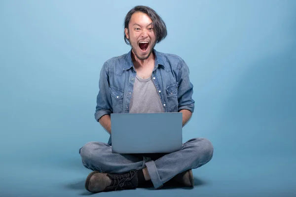 Young bearded man sit down on floor using computer in blue background. Happy Asian young hipster using laptop, full body shot. Young generation hipster working concept.