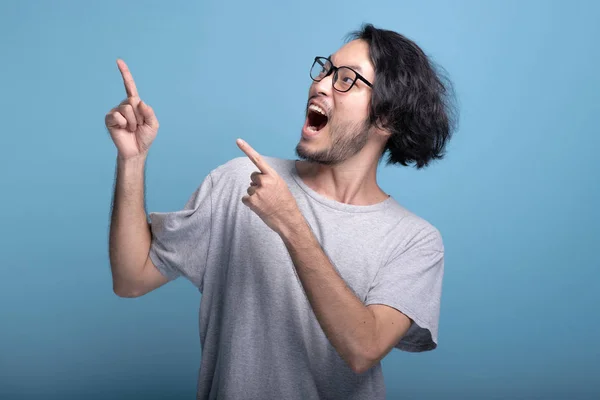 Young bearded man pointing finger in blue background. Excited Asian young hipster pointing finger at copy space, half body shot. Young generation hipster concept.