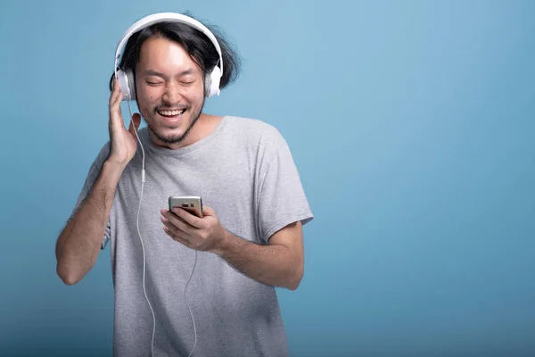 年轻的胡子男子浏览播放列表在手机上的蓝色背景 快乐的亚洲青年时髦使用白色耳机听音乐 半身体射击 年轻一代时髦的概念 — 图库照片