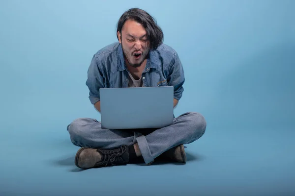 Young bearded man sit down on floor using computer in blue background. Very angry Asian young hipster using laptop, full body shot. Young generation hipster working concept.