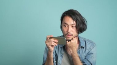 Genç erkek yenilikçi mobil ileti uygulama gelen kötü haber bulmak için seyir onun telefon sakallı. Asya adam cep telefonu arayan sıkılmış ve çok kızgın olarak o kötü haber mavi arka plan ile gördüm.
