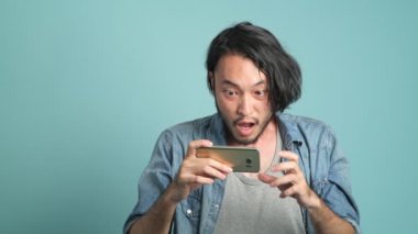 Genç erkek hipster yeni mobil ileti uygulamadan büyük bulmak için seyir onun telefon sakallı. Cep telefonu ve son derece mutlu o gördüğünüz gibi iyi haber mavi arka plan ile sıkılmış Asyalı adam.