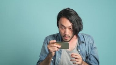 Genç erkek hipster yeni mobil ileti uygulamadan büyük bulmak için seyir onun telefon sakallı. Cep telefonu ve son derece mutlu o gördüğünüz gibi iyi haber mavi arka plan ile sıkılmış Asyalı adam.
