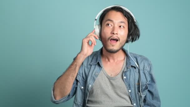 若いひげを生やした男性のヒップな音楽を聴きます 青い背景で彼の音楽を楽しむヘッドフォンを身に着けているアジア人男性 — ストック動画