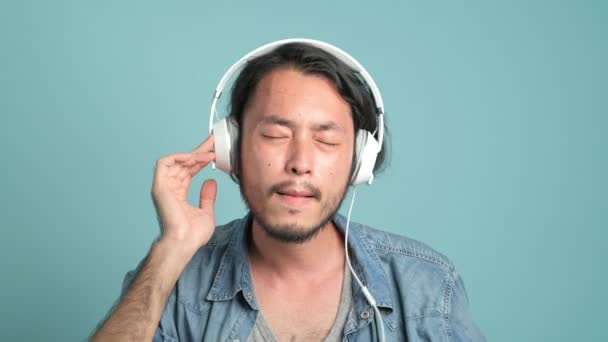 年轻的大胡子男性时髦听音乐 亚洲人佩戴耳机享受他的音乐与蓝色背景 — 图库视频影像