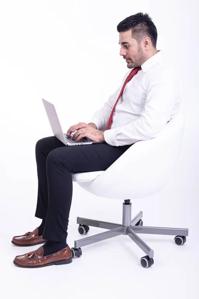 Businessman sitting on white chair isolated. Handsome young indian businessman using laptop portrait, confident looks. Full length side shot.