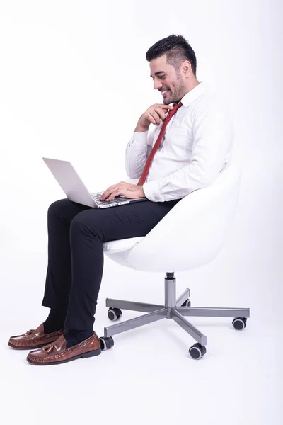 Businessman sitting on white chair isolated. Handsome young indian businessman using laptop portrait, confident smile looks. Full length side shot.