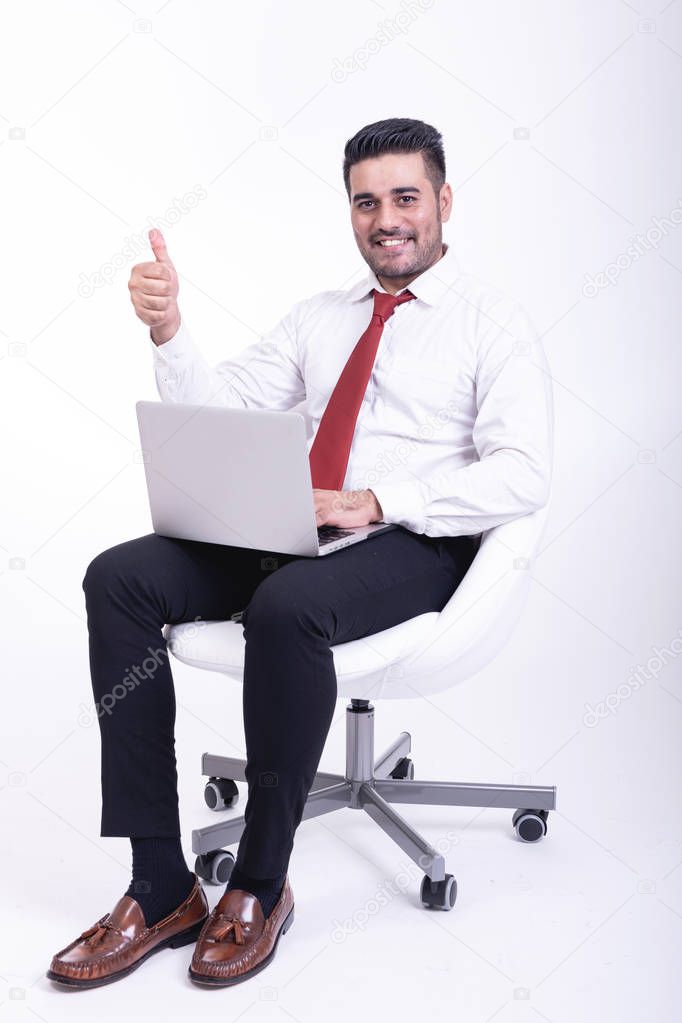 Businessman sitting on white chair isolated. Handsome young indian businessman using laptop portrait, thumb up looks. Full length side shot.