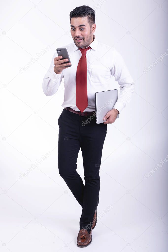 Businessman isolated. Handsome young indian businessman holding laptop, looking at his phone, very surprise looks. Full length shot.