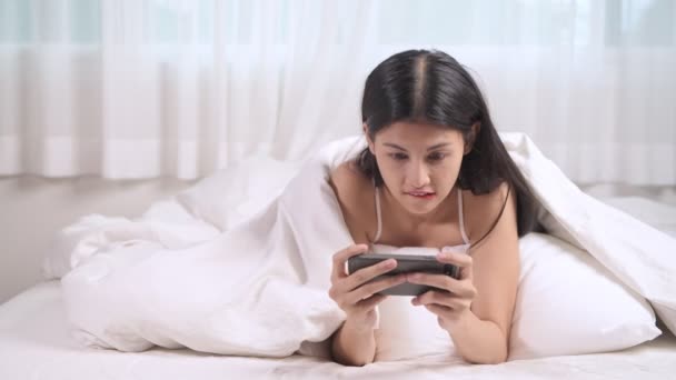 女性がベッドでモバイル ゲームをプレイします アジアの女性がベッドで横になっている携帯電話を使用して — ストック動画