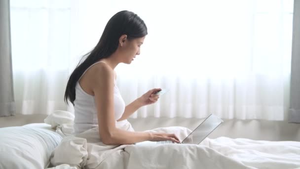 若いアジア女性ベッドに座っているとクレジット カードとノート パソコンにオンライン購入を支払う — ストック動画