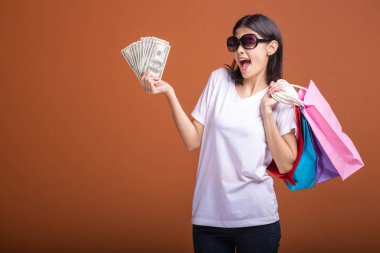 Alışveriş çantası ve turuncu arka planda izole para tutan kadın. Genç Asyalı kadın beyaz tişört içinde çok mutlu ruh hali güneş gözlük. Tatil alışveriş kavramı.