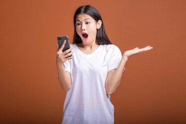 Cep telefonuyla turuncu arka plan üzerinde izole kadın. Asyalı kadın beyaz t-shirt, poz, avuç şok. Hipster yaşam tarzı kavramı.