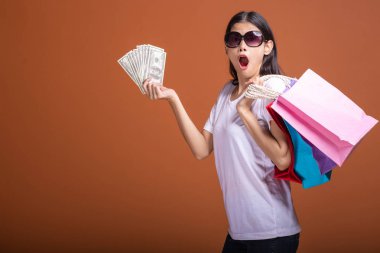 Alışveriş çantası ve turuncu arka planda izole para tutan kadın. Genç Asyalı kadın beyaz tişört içinde çok sürpriz havamda güneş gözlük. Tatil alışveriş kavramı.
