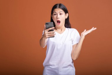 Cep telefonuyla turuncu arka plan üzerinde izole kadın. Asyalı kadın beyaz t-shirt, poz, avuç şok. Hipster yaşam tarzı kavramı.