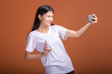 Cep telefonuyla turuncu arka plan üzerinde izole kadın. Asyalı kadın beyaz tişört içinde selfie poz. Hipster yaşam tarzı kavramı.