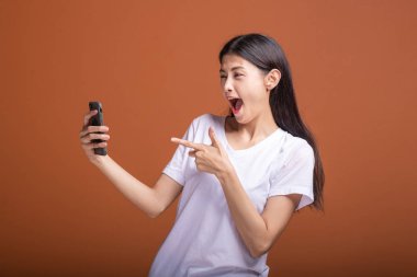 Cep telefonuyla turuncu arka plan üzerinde izole kadın. Asyalı kadın beyaz t-shirt, mutlu poz, telefon noktası parmak. Hipster yaşam tarzı kavramı.