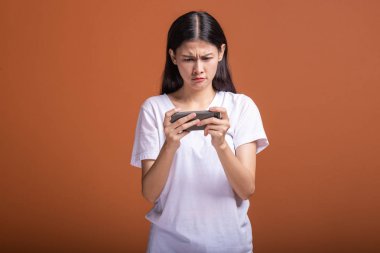 Kadın turuncu arka plan üzerinde izole mobil oyun oynuyor. Beyaz t-shirt, Genç Asyalı kadın üzgün ruh hali. Genç hipster yaşam tarzı kavramı.