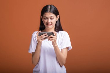 Kadın turuncu arka plan üzerinde izole mobil oyun oynuyor. Beyaz t-shirt, odak ruh hali içinde Genç Asyalı kadın. Genç hipster yaşam tarzı kavramı.