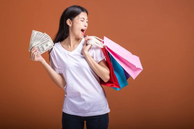 Alışveriş çantası ve turuncu arka planda izole para tutan kadın. Beyaz tişört çok heyecanlı ruh hali içinde Genç Asyalı kadın. Tatil alışveriş kavramı.