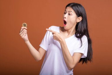 Turuncu arka planda izole bit sikke tutan kadın. Genç Asyalı kadın kazanmak ruh şok edici içinde beyaz tişört içinde. Genç zengin hipster online ticaret kavramı.