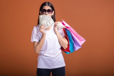 Alışveriş çantası ve turuncu arka planda izole para tutan kadın. Genç Asyalı kadın beyaz tişört içinde mutlu ruh hali güneş gözlük. Tatil alışveriş kavramı.