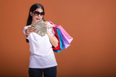 Alışveriş çantası ve turuncu arka planda izole para tutan kadın. Genç Asyalı kadın beyaz tişört içinde mutlu ruh hali güneş gözlük. Tatil alışveriş kavramı.