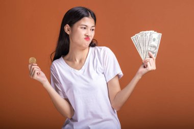 Turuncu arka planda izole bit sikke tutan kadın. Genç Asyalı kadın çok üzgün ruh hali, beyaz t-shirt dolar olarak seyir içinde. Genç zengin hipster online ticaret kavramı.