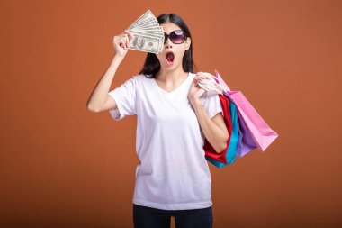 Alışveriş çantası ve turuncu arka planda izole para tutan kadın. Genç Asyalı kadın beyaz tişört içinde çok sürpriz havamda güneş gözlük. Tatil alışveriş kavramı.