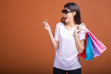 Alışveriş çantası ve turuncu arka planda izole bitcoin tutan kadın. Genç Asyalı kadın beyaz tişört içinde heyecan havamda güneş gözlük. Online para birimi alışveriş kavramı.