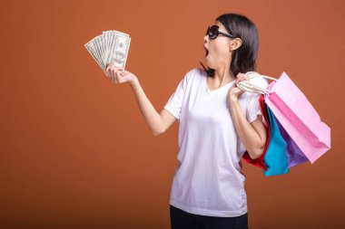 Alışveriş çantası ve turuncu arka planda izole para tutan kadın. Beyaz t-shirt, güneş gözlüğü son derece heyecanlı ruh hali içinde Genç Asyalı kadın. Tatil alışveriş kavramı.