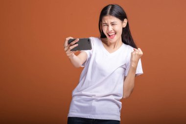 Cep telefonuyla turuncu arka plan üzerinde izole kadın. Asyalı kadın beyaz t-shirt, büyük kazanç poz içinde. Hipster yaşam tarzı kavramı.