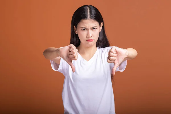 Upset woman isolated over orange background. Asian woman in white t-shirt doing thumb down pose, upset mood.