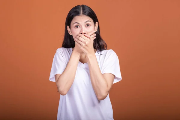 Surprise woman over orange background. Young Asian hipster woman in white t-shirt with surprise pose, two hands cover mouth. Young woman hipster concept.