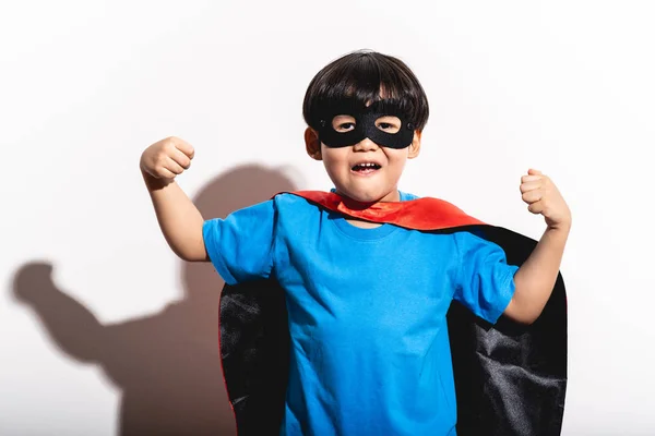 Young boy super hero portrait in white background with hard light. Mixed race boy in blue shirt, jean, mask, cape. Super strength pose.