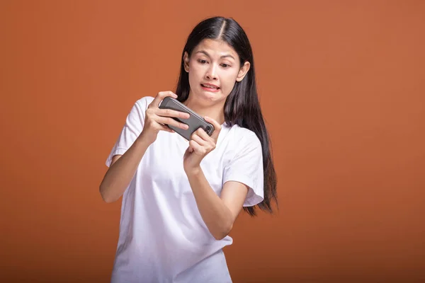Woman playing mobile game isolated over orange background. Young asian woman in white t-shirt, extremely focus mood. Young hipster lifestyle concept.