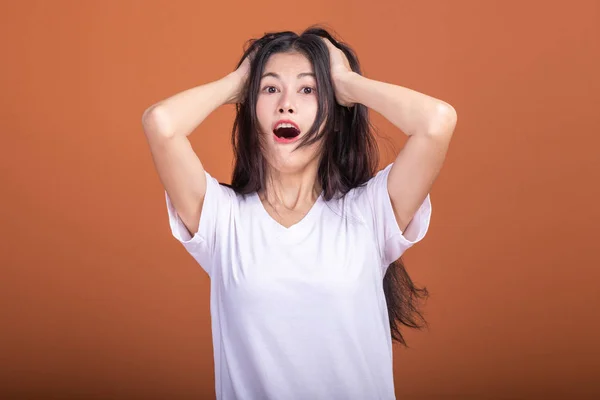 Surprise woman over orange background. Young Asian hipster woman in white t-shirt with surprise pose, pulling hair. Young woman hipster concept.