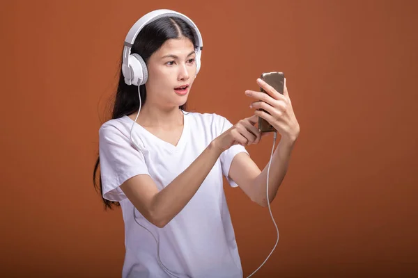 在橙色背景下 通过电话隔离的妇女听音乐 年轻的亚洲女人用手机听音乐 浏览音乐 时髦的生活方式概念 — 图库照片