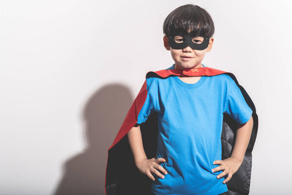 Young boy super hero portrait in white background with hard light. Mixed race boy in blue shirt, jean, mask, cape. Ready to fight pose.