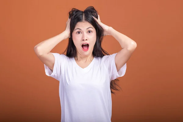 Surprise woman over orange background. Young Asian hipster woman in white t-shirt with surprise pose, pulling hair. Young woman hipster concept.