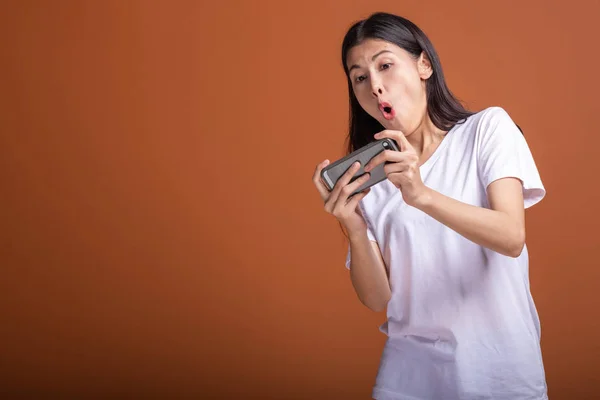Woman playing mobile game isolated over orange background. Young asian woman in white t-shirt, shock mood. Young hipster lifestyle concept.