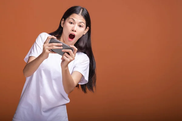 Woman playing mobile game isolated over orange background. Young asian woman in white t-shirt, shock mood. Young hipster lifestyle concept.