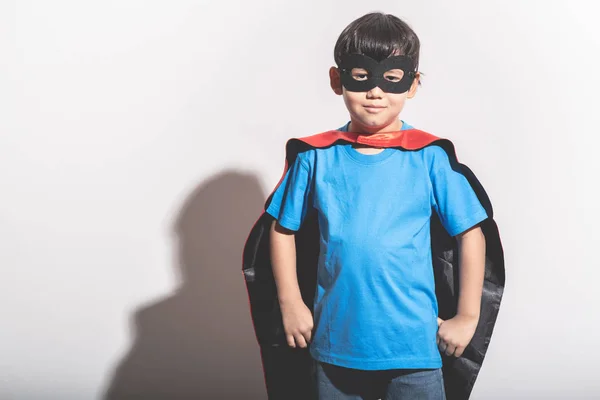 Young boy super hero portrait in white background with hard light. Mixed race boy in blue shirt, jean, mask, cape. Ready to fight pose.